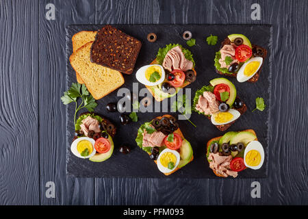 Thunfisch Sandwiches mit Avocado, Salat, Tomaten, schwarze Oliven und hart gekochte Ei auf ein Roggen und Mais geröstetes Brot auf einem schwarzen Schiefer Fach Stockfoto