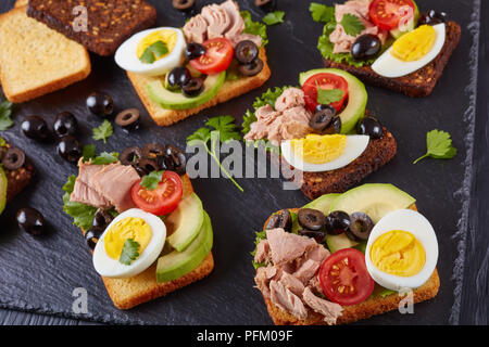 Scheiben Thunfisch Sandwiches mit Avocado, Salat, Tomaten, schwarze Oliven und hart gekochte Ei auf Roggen und Mais getoasteten Brotscheiben auf schwarzem Schiefer board o Stockfoto