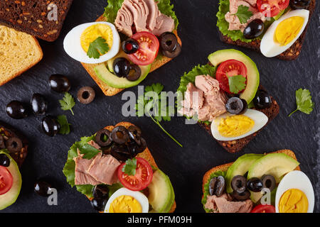 In der Nähe von Thun Sandwiches mit Avocado, Salat, Tomaten, schwarze Oliven und hart gekochte Ei auf Roggen und Mais getoasteten Brotscheiben auf einem schwarzen s Stockfoto