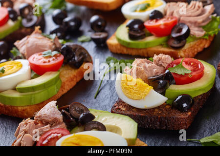 Toast mit Thunfisch in Dosen, Avocado, Salat, Tomaten, schwarze Oliven und hart gekochte Ei auf Roggen und Mais getoasteten Brotscheiben auf schwarzem Schiefer boa Stockfoto