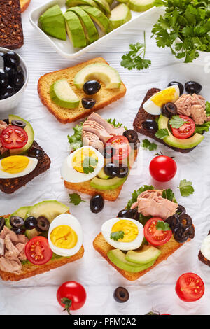 Toast mit Thunfisch in Dosen, Avocado, Salat, Tomaten, schwarze Oliven und hart gekochte Ei auf Roggen und Mais getoasteten Brotscheiben auf einem Papier, vertikal Stockfoto