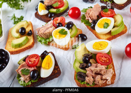 Toast mit Thunfisch in Dosen, Avocado, Salat, Tomaten, schwarze Oliven und hart gekochte Ei auf Roggen und Mais getoasteten Brotscheiben auf einem weißen Holz- ta Stockfoto