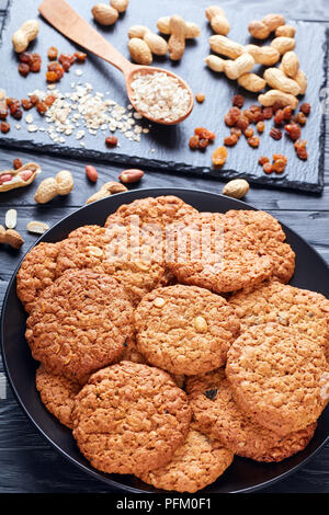 Nahaufnahme der leckere Haferflocken Rosinen und peanut Cookies auf eine schwarze Platte mit Zutaten auf einer Schiefertafel Fach im Hintergrund, vertikale Ansicht von oben Stockfoto