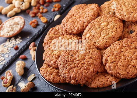Nahaufnahme der leckere Haferflocken Rosinen und peanut Cookies auf eine schwarze Platte mit Zutaten auf einer Schiefertafel Fach im Hintergrund, horizontale Ansicht von abov Stockfoto