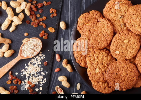 Nahaufnahme der leckere Haferflocken Rosinen und peanut Cookies auf eine schwarze Platte mit Zutaten auf einer Schiefertafel Fach im Hintergrund, horizontale Ansicht von abov Stockfoto