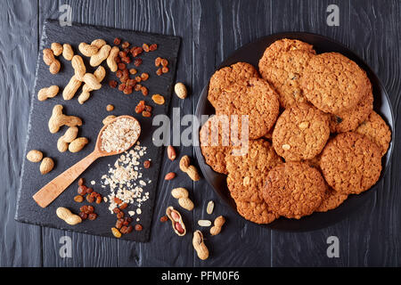 Leckere Haferflocken Rosinen und peanut Cookies auf eine schwarze Platte mit Zutaten auf einer Schiefertafel Fach im Hintergrund, horizontale Ansicht von oben, flach Stockfoto