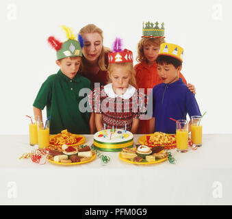 Mädchen bläst die Kerzen auf der Geburtstagstorte, die von Kindern und Erwachsenen umgeben, Kinder tragen Papier Kronen Stockfoto