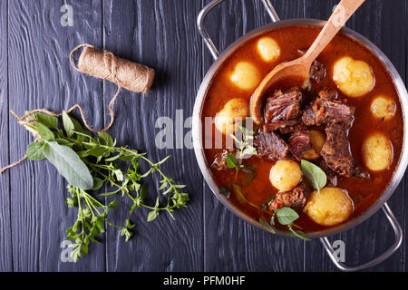 Rinderragout mit zarten Cubes von Fleisch, neue Kartoffeln, Möhren und Kräuter in einem Metall Topf auf schwarzer Holztisch mit Blumenstrauß aus aromatischen Kräutern, Aussicht f Stockfoto