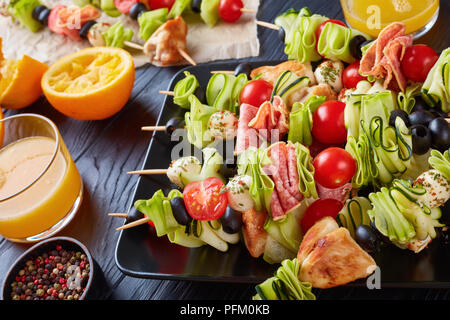 Spiesse mit Hähnchenfleisch, Zucchini, Tomaten, Mozzarella Kugeln, Scheiben Salami, Oliven auf einer schwarzen Platte auf einem Holztisch mit Orangensaft in Glas c Stockfoto