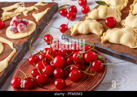 Ungekocht cherry Knödel auf einer hölzernen Schneidebrett mit Teig, Platte mit frischen Kirschen im Vordergrund, close-up Stockfoto