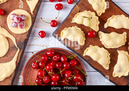 Ungekocht cherry Knödel auf einer hölzernen Schneidebrett mit Teig und die Platte mit frischen Kirschen auf einem weißen rustikale Tabelle, Ansicht von oben, Nahaufnahme, flach Stockfoto