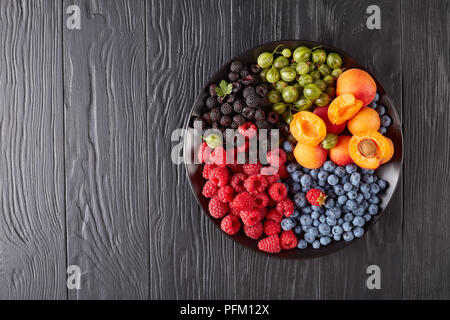 Obst und Beeren Teller mit frischen Bioprodukten Stachelbeeren, Rote und Schwarze Himbeere, Heidelbeere, Aprikose Schichten auf einem schwarzen Holztisch, horizontale Ansicht Stockfoto