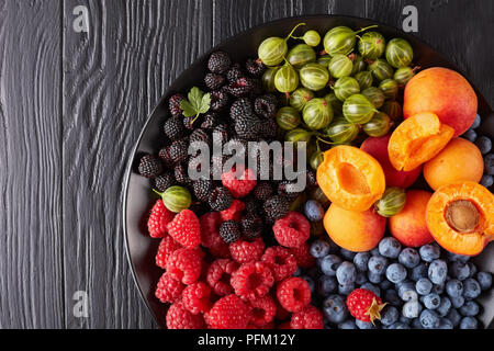 Obst und Beeren Teller mit frischen Bioprodukten Stachelbeeren, Rote und Schwarze Himbeere, Heidelbeere, Aprikose Schichten auf einer schwarzen Platte auf einem Holztisch, horizo Stockfoto