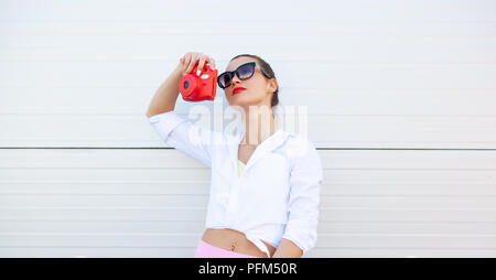 Mode Portrait von hübsch lächelnde Frau mit Sonnenbrille, Foto, das durch die Kamera gegen die graue Wand. Kabellose Kopfhörer, fitness Armband auf der Hand Stockfoto