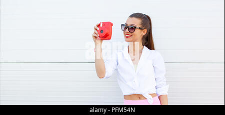 Mode Portrait von hübsch lächelnde Frau mit Sonnenbrille, Foto, das durch die Kamera gegen die graue Wand. Kabellose Kopfhörer, fitness Armband auf der Hand Stockfoto