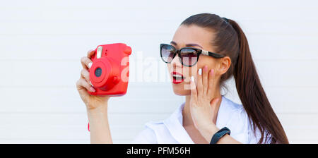 Mode Portrait von hübsch lächelnde Frau mit Sonnenbrille, Foto, das durch die Kamera gegen die graue Wand. Kabellose Kopfhörer, fitness Armband auf der Hand Stockfoto