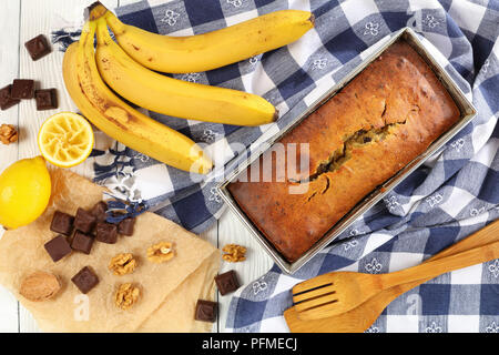 Frisch gebackene leckere Bananen Brot mit Nüssen und Schokolade Stücke in eine Kastenform mit Küchentuch, reife Bananen und Zutaten auf hölzernen Tisch Stockfoto