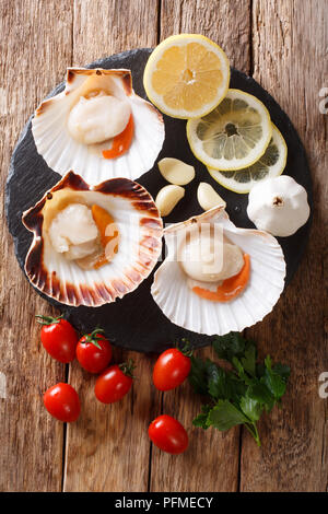 Produkte zum Kochen: raw Jakobsmuscheln in einer Shell und Tomaten, Knoblauch, Petersilie und Zitrone close-up auf einem Holztisch. Senkrechte Draufsicht von oben Stockfoto