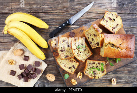 Frisch gebackene leckere Bananen Brot mit Nüssen und Schokolade Stücke in Scheiben schneiden auf Schneidebrett. Zutaten auf rustikalen Holztisch, Ansicht von eine