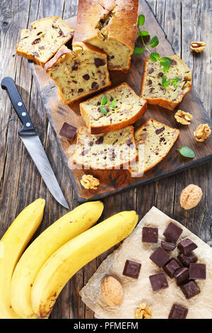 Frisch gebackene leckere Bananen Brot mit Nüssen und Schokolade Stücke in Scheiben schneiden auf Schneidebrett. Zutaten auf rustikalen Holztisch, vertikale vi. Stockfoto