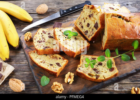 Frisch gebackene leckere Bananen Brot mit Nüssen und Schokolade Stücke in Scheiben schneiden auf Schneidebrett. Zutaten auf rustikalen Holztisch, amerikanische Cu Stockfoto