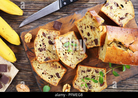 Frisch gebackene leckere Bananen Brot mit Nüssen und Schokolade Stücke in Scheiben schneiden auf Schneidebrett. Zutaten und Messer auf dunklen Holzmöbeln im Landhausstil tab Stockfoto