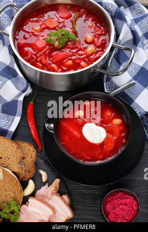 Köstliche rote Beete rote Suppe mit weißen Bohnen oder Borschtsch in Edelstahl Kasserolle schwenken und in Schwarz Schüssel mit Roggenbrot und geräuchertem Speck auf Tisch, Stockfoto