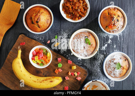 Leckere frisch gebackene selbstgemachte Banane Mutter raisin Muffins in Keramik Förmchen bestreut mit Puderzucker auf schwarzer Holztisch mit Zutaten Stockfoto