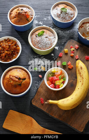 Leckere frisch gebackene selbstgemachte Banane Mutter raisin Muffins in Keramik Förmchen bestreut mit Puderzucker auf schwarzer Holztisch mit Zutaten Stockfoto