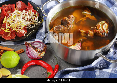 Heiße Brühe Rindfleisch mit Knochen und andere Zutaten für traditionelle vietnamesische Suppe Pho Bo mit rohem Rindfleisch, Sojasprossen, Gewürze und Grüns, Ansicht von oben, Clos Stockfoto