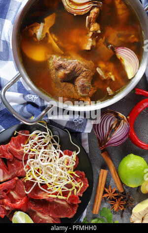 Heiße Brühe Rindfleisch mit Knochen und andere Zutaten für traditionelle vietnamesische Suppe Pho Bo mit rohem Rindfleisch, Sojasprossen, Gewürze und Grüns, vertikale Ansicht von ab Stockfoto