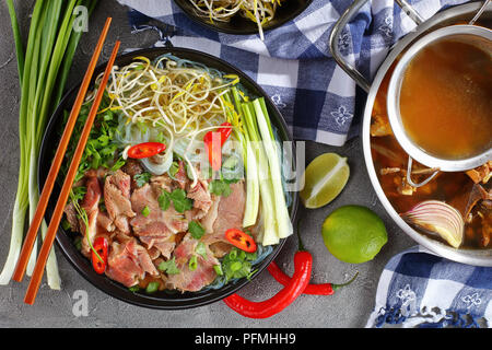 Nahaufnahme der köstliche vietnamesische Suppe Pho Bo mit rohen Rinderfilet Rindfleisch Scheiben in schwarz Schüssel mit Stäbchen. hot Rindfleisch Knochen Brühe in Edelstahl Pfanne und oth Stockfoto