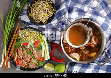 Köstliche vietnamesische Suppe Pho Bo in schwarz Schüssel mit Stäbchen. hot Rindfleisch mit Knochen Brühe in Edelstahl Pfanne und andere Zutaten auf Tabelle, Ansicht von oben Stockfoto