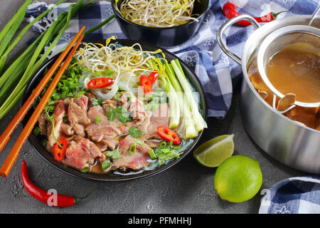 Vietnamesische Suppe Pho Bo mit Reis, Nudel, roh Rumpsteak Rindfleisch Scheiben, Sojasprossen in schwarz Schüssel mit Stäbchen. hot Rindfleisch Knochen Brühe in Edelstahl Pfanne ein Stockfoto