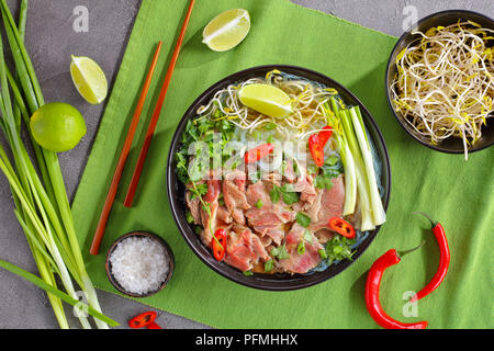 Vietnamesische Suppe Pho Bo mit Reis, Nudel, roh Rumpsteak Rindfleisch Scheiben, Sojasprossen, Kalk, Frühlingszwiebeln in schwarz Schüssel mit Stäbchen, Peperoni auf Grün Stockfoto