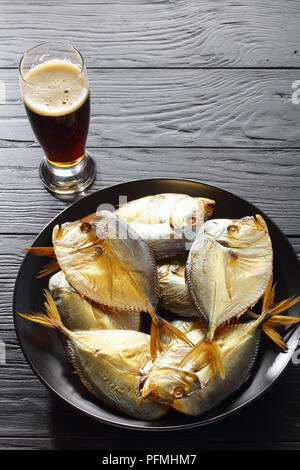 Köstlich kalt geräuchert Mond Fisch auf schwarzem Teller und Glas von geschäumten dunkles Bier auf schwarzem Holz- Tabelle, Ansicht von oben Stockfoto