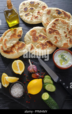 Gebratene hausgemachte heiße pita Brot auf schwarzen Holztisch, frische tzatziki Sauce in der Schüssel und Zutaten auf Stein Schneidbrett, vertikale Ansicht von abov Stockfoto