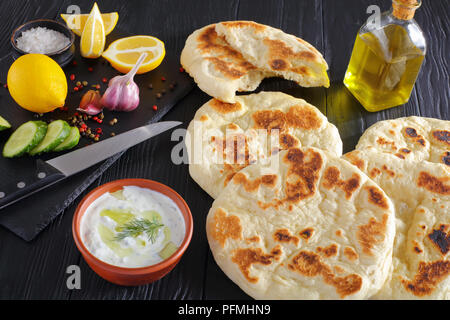 Frisch gebackenes warmes Fladenbrot auf schwarzen Holztisch, frische tzatziki Sauce in der Schüssel und Zutaten auf Stein Schneidbrett, Ansicht von oben, close-up Stockfoto