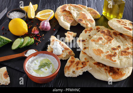 Frisch gebackenes Fladenbrot auf schwarzer Holztisch mit herzhaften tzatziki Soße Zutaten auf Stein Schneidbrett, horizontale Ansicht von oben, close-up Stockfoto