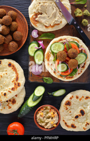 Leckeren Falafels auf pan fried Fladenbrot mit würziger Hummus, Tomaten und Gurkenscheiben. Zutaten auf schwarzen Holztisch, horizontale Ansicht von abov Stockfoto