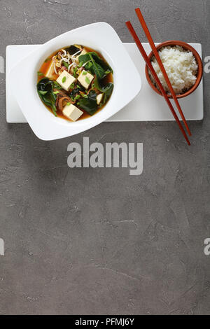 Miso Suppe serviert in einer weißen Schüssel mit Essstäbchen und gedämpftem Reis auf weißer Keramik Tablett auf konkreten Hintergrund, vertikale Ansicht von oben Stockfoto