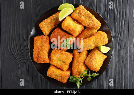 Köstliches Stück weißen Fisch paniert mit Kokos und Brotkrumen auf schwarze Platte mit Kalk, Scheiben, auf Schwarz Holz- Tabelle, Ansicht von oben Stockfoto