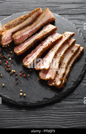 Lecker würzig geräucherter Speck in Scheiben geschnitten auf Schwarze runde Steinplatte auf Holztisch, vertikale Ansicht von oben Stockfoto