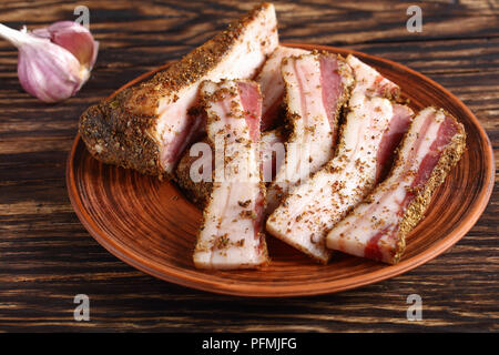 Nahaufnahme der köstliche würzige Luft getrocknet in Scheiben Speck auf Lehm Platte auf rustikalen Holztisch mit Knoblauch im Hintergrund, horizontale Ansicht von oben Stockfoto