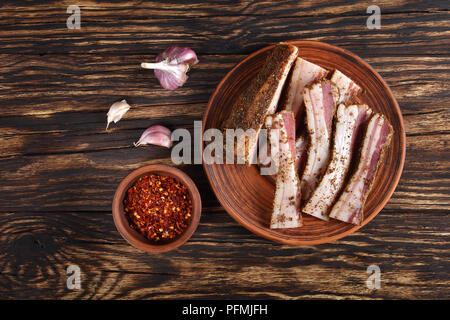 Leckere würzige Luft Speck in Scheiben geschnitten auf Lehm Platte auf rustikalen Holztisch mit Knoblauch und Chili Flocken in die Schüssel am Hintergrund getrocknet, horizontale Ansicht aus Stockfoto