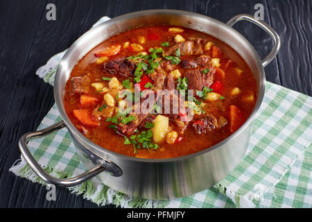 Leckere heiße Ungarisches Gulasch mit Rindfleisch, Paprika, Gemüse und Teigwaren csipetke - kleine Ei in Edelstahl Topf auf schwarzen Holztisch, Klasse Stockfoto