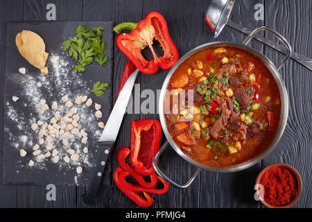 Leckere heiße Ungarisches Gulasch mit Rindfleisch, Paprika, Gemüse, in einen Topf. raw csipetke mit Teig an Bord, Paprika und suppenkelle am Tisch, Stockfoto