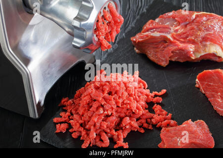 Elektrischer Fleischwolf macht forcemeat von frischem Rindfleisch auf schwarzem Schiefer Platte auf einem hölzernen Hintergrund, Ansicht von oben, close-up Stockfoto