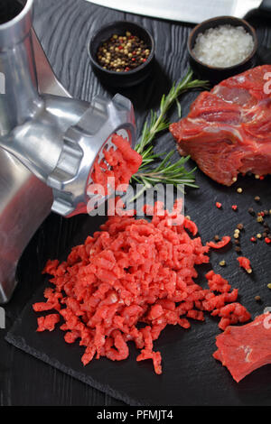 Elektrischer Fleischwolf macht forcemeat von frischem Rindfleisch auf schwarzem Schiefer Platte auf einem Holztisch mit Messer, Pfeffer, Chili und frischem Rosmarin, Stockfoto