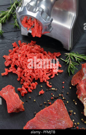 Fleischwolf macht frisches Rindfleisch forcemeat auf schwarzem Schiefer Platte auf Holztisch mit Pfeffer und frischem Rosmarin im Hintergrund, Blick von oben, in der Nähe - u Stockfoto
